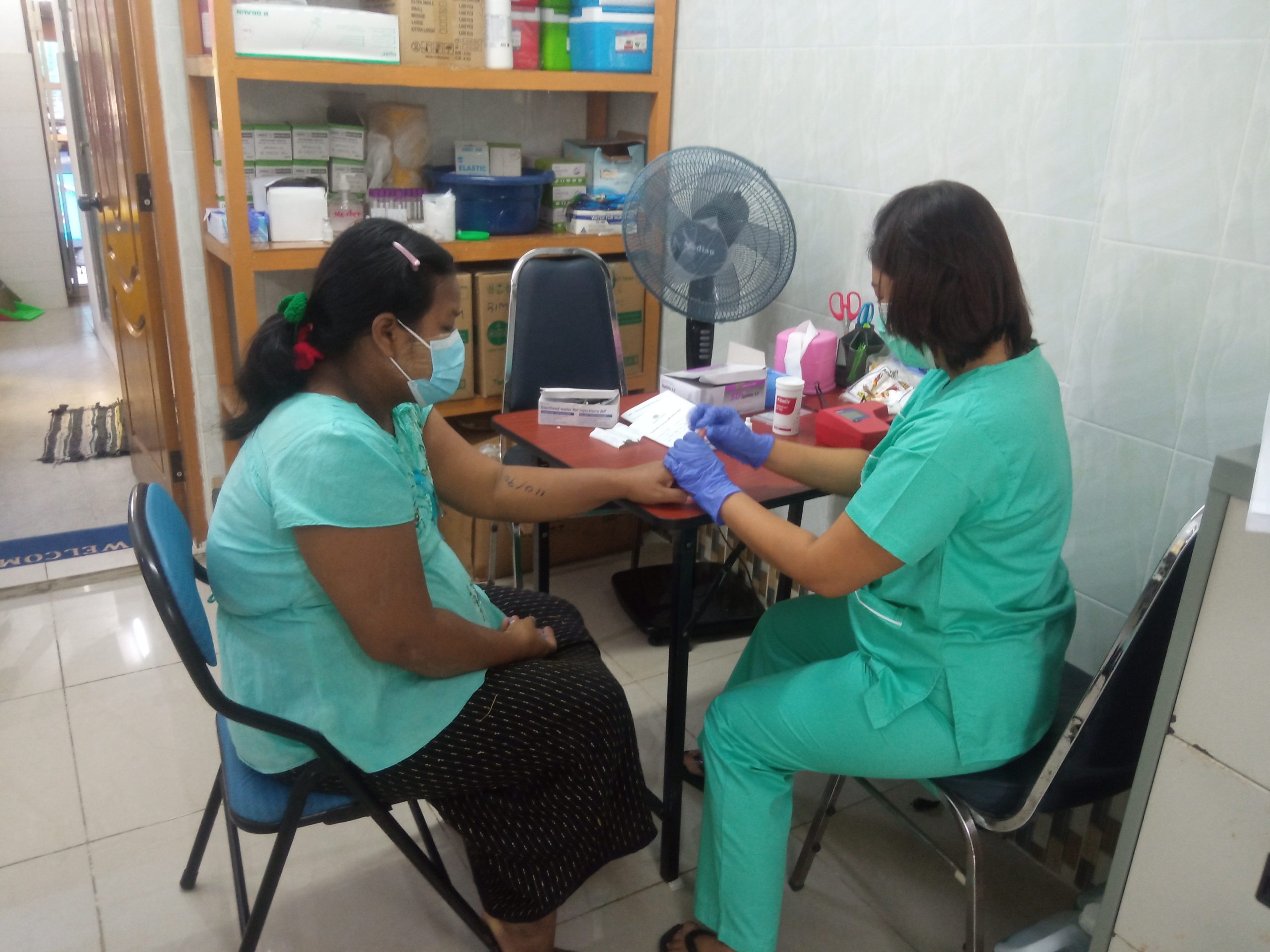 Seated women in a green has a finger prick test taken by a seated nurse who is also in greentop