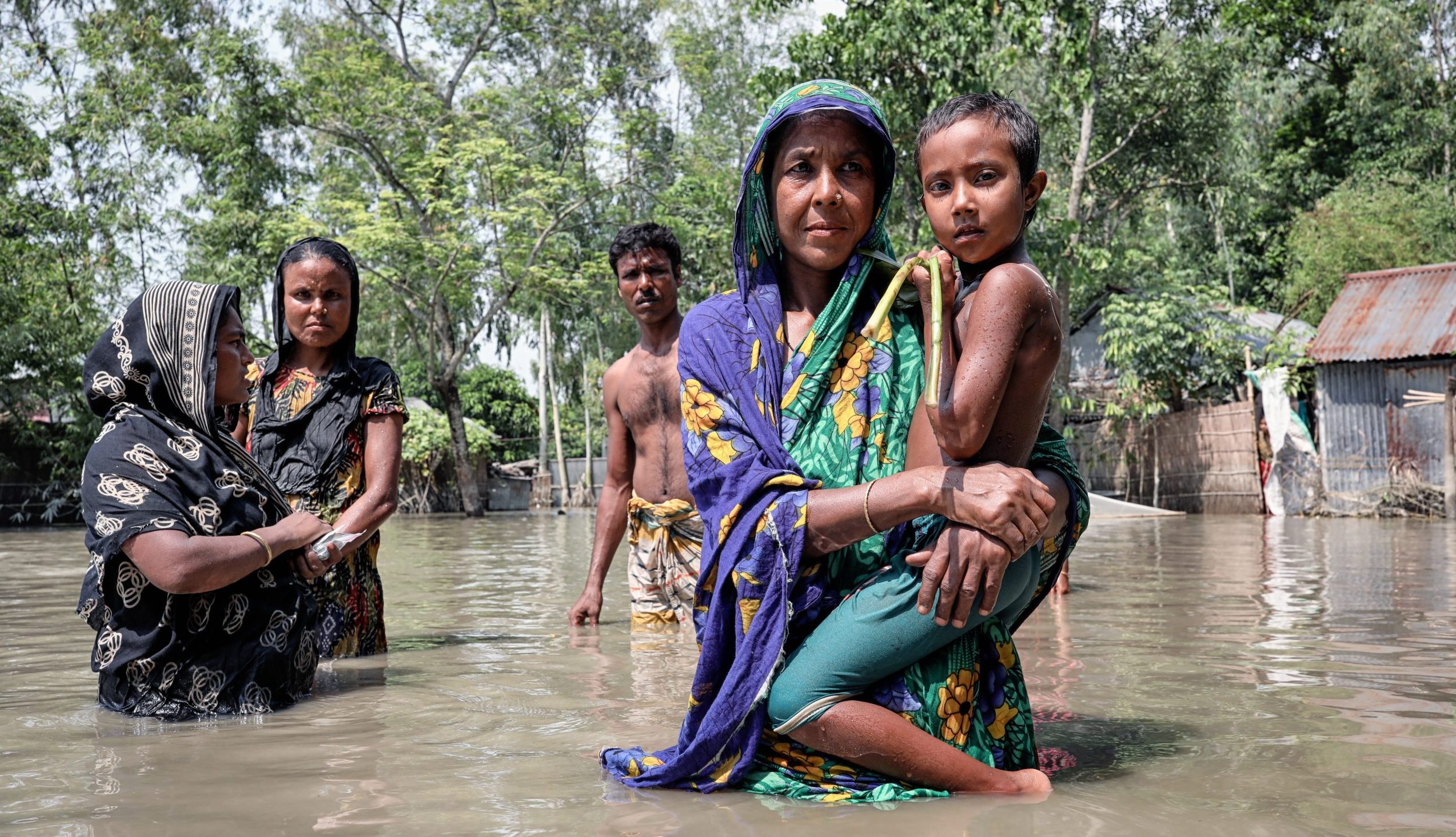 A Bangladeshi woman holds a child while standing in brown flood water. Behind are two women and a man.